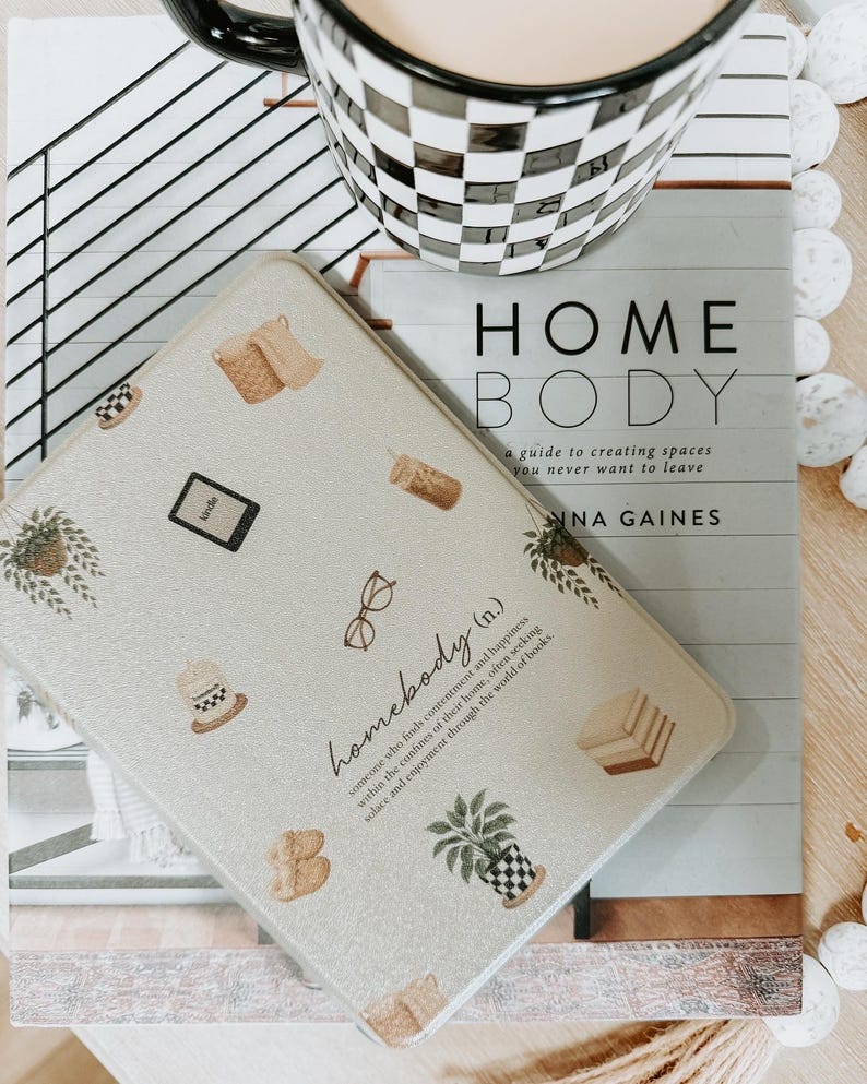 May include: A decorative, light-colored tray with various illustrations, including a potted plant, glasses, and a coffee cup. The tray is on top of a book titled "HOME BODY" and a black and white checkered mug.