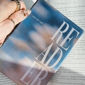 May include: A rectangular reader bookmark with a gradient blue and peach background. The word "READER" is printed in large white letters, with "LET'S GET LOST" above. The bookmark is held over an open book.