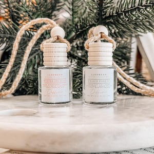 May include: Two glass diffuser bottles with wooden tops and rope hangers. One bottle reads "Winter Orchard" and the other "Fraser Forest." The bottles sit on a white marble surface, with a blurred background of greenery.