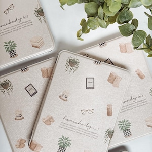 May include: Beige Kindle covers with a repeating pattern of potted plants, books, eyeglasses, and coffee cups. The word "homebody" is printed on the cover. Green leaves are in the background.