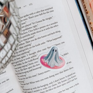 May include: A sticker of a gray ghost floating on a pink inflatable ring is placed on an open book. The book's pages are filled with black text. The sticker has a cute, cartoonish design, and the book is open to page 173.