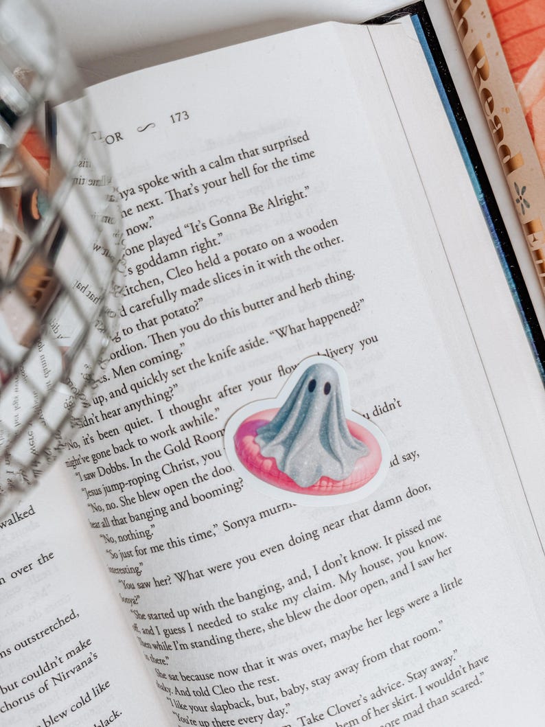 May include: A sticker of a gray ghost floating on a pink inflatable ring is placed on an open book. The book's pages are filled with black text. The sticker has a cute, cartoonish design, and the book is open to page 173.