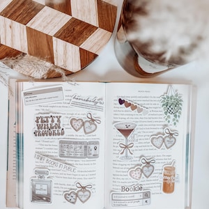 May include: An open book with decorative stickers, including heart-shaped tags, a cocktail, and a coffee drink. The stickers have text like "Petty When Provoked" and "I Sniff Books in Public." A wooden cutting board and a glass vase are in the background.
