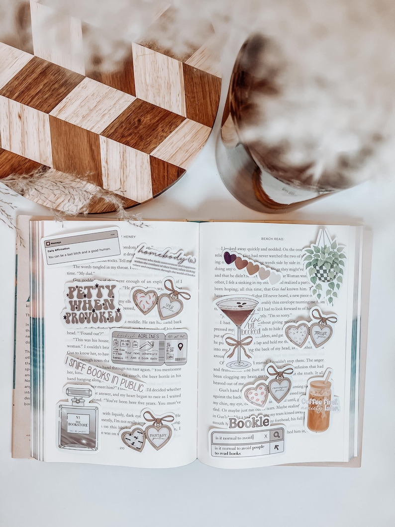 May include: An open book with decorative stickers, including heart-shaped tags, a cocktail, and a coffee drink. The stickers have text like "Petty When Provoked" and "I Sniff Books in Public." A wooden cutting board and a glass vase are in the background.