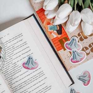 May include: A still life featuring a book with a decorative bookmark, white tulips, and several ghost-shaped stickers. The stickers have a gray ghost design with a pink inner tube. The book is open to page 173.