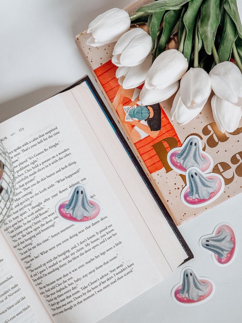 May include: A still life featuring a book with a decorative bookmark, white tulips, and several ghost-shaped stickers. The stickers have a gray ghost design with a pink inner tube. The book is open to page 173.