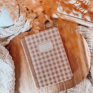 May include: A Composition Notebook with a brown and white gingham pattern, labeled "Limited Edition," rests on a wooden cutting board. The notebook is surrounded by a beige knitted blanket and dried plants. The words "Let's get" are visible in the background.