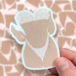 May include: A sticker featuring a stylized illustration of a person in a bikini. The bikini top and bottom are light pink with a white daisy pattern. The person's skin tone is light brown. The sticker is held by a hand.