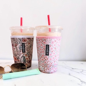 May include: Two clear plastic iced coffee cups with red straws, each encased in a fabric sleeve featuring the brand name LLOUBEAN. One sleeve has a dense wildflower print, while the other displays a pink floral pattern with ribbon bows.