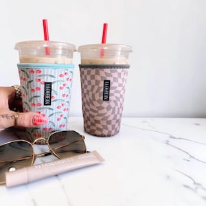 May include: Two clear plastic iced coffee cups with red straws, each inside a fabric sleeve labeled LLOUBEAN. One sleeve features a light blue and white striped pattern with red cherries, while the other has a tan and brown wavy checkerboard print.