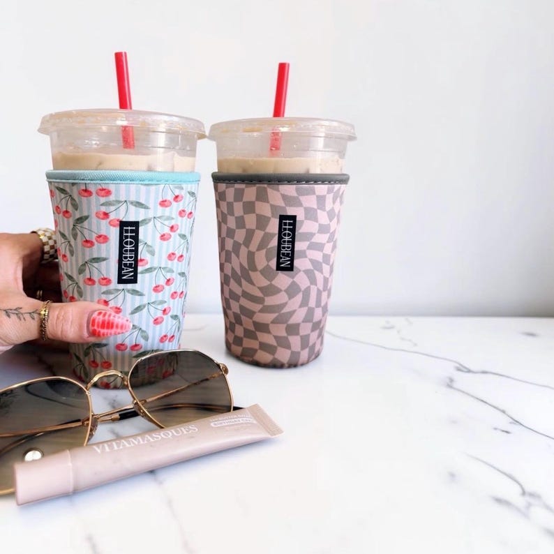 May include: Two clear plastic iced coffee cups with red straws, each inside a fabric sleeve labeled LLOUBEAN. One sleeve features a light blue and white striped pattern with red cherries, while the other has a tan and brown wavy checkerboard print.