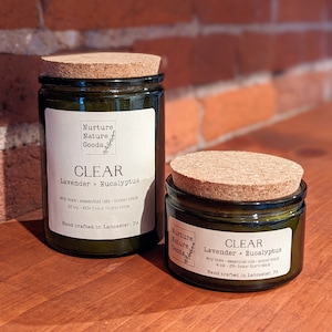May include: Two glass jars of soy wax candles with cork lids. The labels read "CLEAR Lavender + Eucalyptus" and "Nurture Nature Goods." The larger candle is 12 oz with a 60+ hour burn time, and the smaller is 4 oz with a 20+ hour burn time.