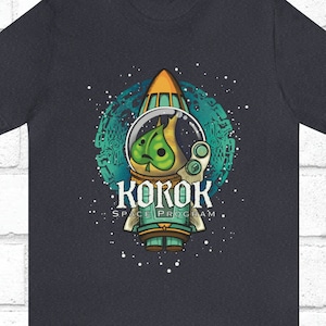 May include: Dark grey t-shirt with a green and brown cartoon character in a rocket ship. The character is wearing a space helmet and the rocket ship is surrounded by stars. The text "KOROK SPACE PROGRAM" is printed below the rocket ship.