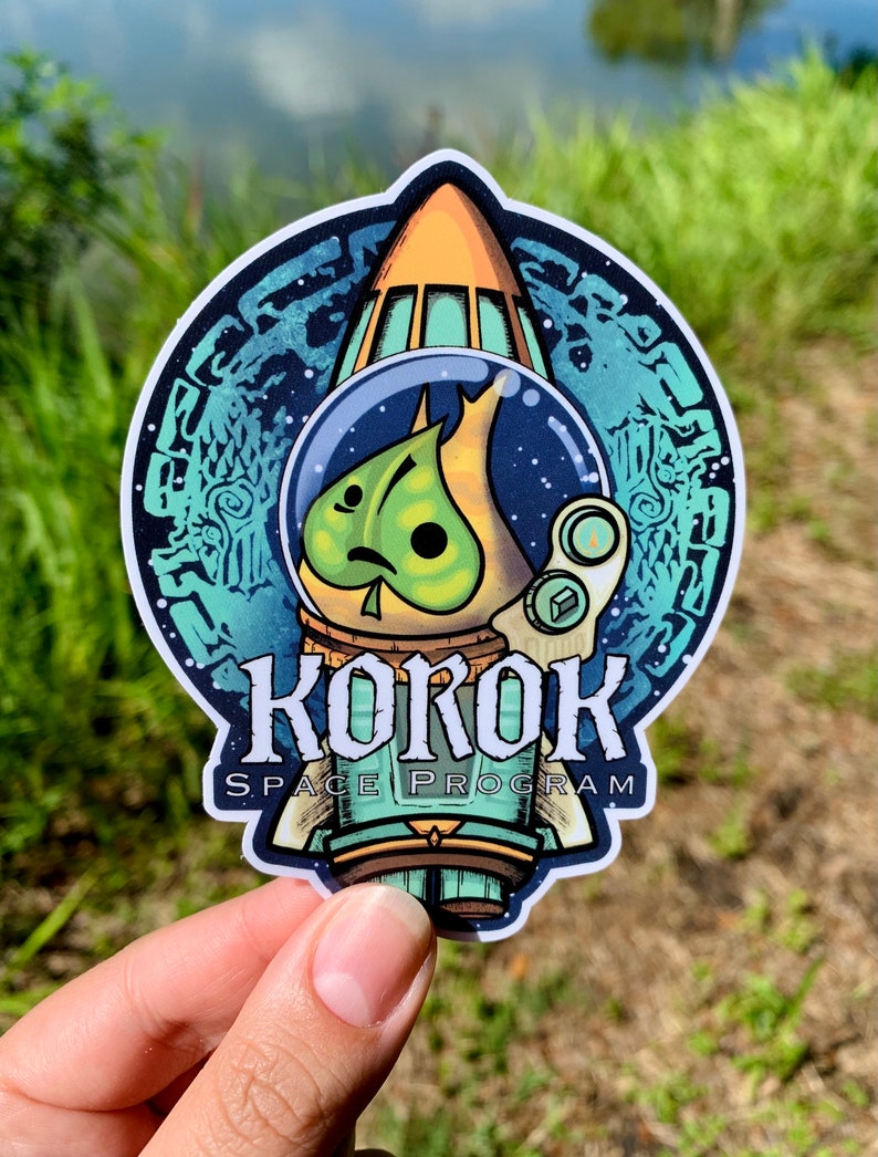 Puede incluir: Una pegatina redonda con un personaje Korok verde de la serie de videojuegos The Legend of Zelda. El Korok lleva un casco espacial y est&aacute; dentro de una nave espacial. La pegatina tiene el texto "Korok Space Program" alrededor de la parte inferior.