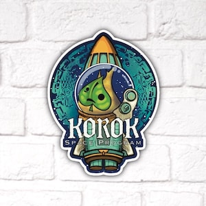 May include: A green, cartoon character wearing a space suit and helmet, inside a rocket ship. The rocket ship is surrounded by a blue and green circle with a pattern of stars and swirls. The text "KOKOK SPACE PROGRAM" is below the rocket ship.