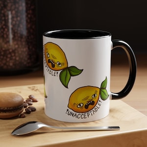 May include: White ceramic mug with a black handle and rim. The mug features a repeated design of yellow lemons with green leaves and the text "UNACCEPTABLE!" printed on the mug.