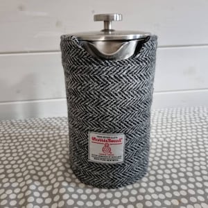 Op de afbeelding: Een roestvrijstalen French press koffiezetapparaat met een zwarte en witte tweed stoffen hoes. De hoes heeft de tekst "Harris Tweed" en "Hand Woven Cloth" erop gedrukt.
