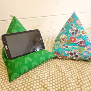 Puede incluir: Dos almohadas triangulares de tela con diferentes patrones. Una almohada es verde con un patrón de arcoíris y sostiene una tableta. La otra almohada es azul con un patrón de calavera y flores.