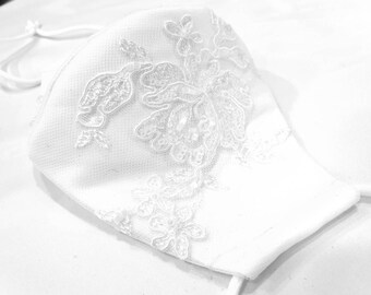 May include: White face mask with delicate lace detailing. The lace features a floral design with small pearl-like embellishments.