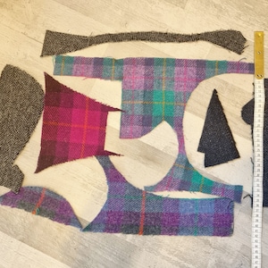 Op de afbeelding: Een assortiment van stoffenresten in verschillende kleuren en patronen, waaronder ruit, herringbone en tweed. De stoffenresten liggen op een houten oppervlak met een meetlint.