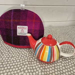 Op de afbeelding: Een rood-wit gestreepte theepot met een kleurrijk ontwerp. De theepot staat op een grijs-wit geblokt oppervlak. Een paars-rode geruite theepotwarmer is op de achtergrond te zien.