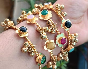 May include: Gold-toned bangles with floral and bell accents. Each bangle features a different colored gemstone, including pink, green, black, and red. The bangles are intricately designed with twisted metal and small, dangling bells.