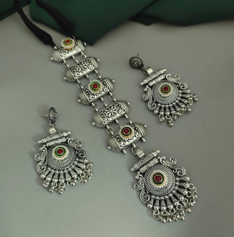 Puede incluir: Un conjunto de collar y pendientes plateados. El collar presenta eslabones decorativos en forma de barril con detalles florales y detalles circulares rojos y verdes. Los pendientes coinciden con el collar, con cuentas plateadas colgantes.