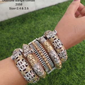 May include: A collection of silver and gold-toned bangles with floral and geometric designs. The bangles are worn on a wrist, showcasing a variety of styles, including engraved and beaded patterns. The text "Dualtone Bangle combo 2058 Size=2.4 & 2.6" is visible.
