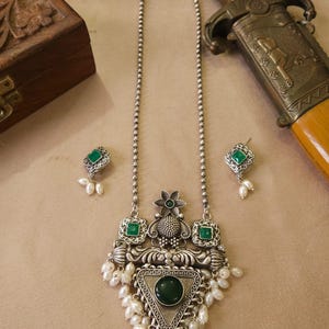Puede incluir: Un conjunto de collar y pendientes plateados. El collar presenta un colgante con una piedra verde grande, piedras verdes m&aacute;s peque&ntilde;as y perlas blancas. Los pendientes tienen piedras verdes y perlas a juego. Una daga decorativa est&aacute; en el fondo.