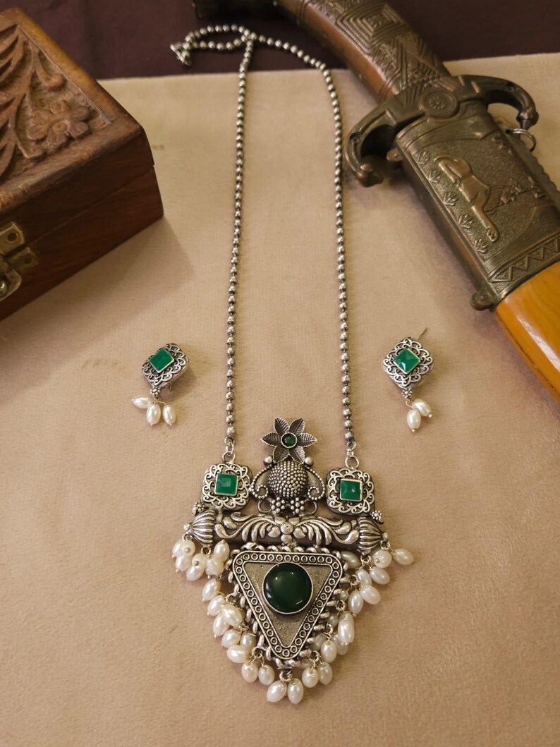 Puede incluir: Un conjunto de collar y pendientes plateados. El collar presenta un colgante con una piedra verde grande, piedras verdes m&aacute;s peque&ntilde;as y perlas blancas. Los pendientes tienen piedras verdes y perlas a juego. Una daga decorativa est&aacute; en el fondo.