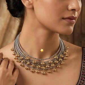 以下が含まれることがあります： シルバーのバンドとゴールドのベルチャームが付いた、マルチストランドネックレス。 ネックレスは首の周りに着用し、ベルチャームが垂れ下がっています。 このジュエリーは、特別な機会に適したステートメントピースです。