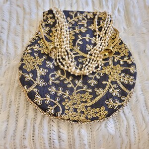 May include: A round, black drawstring bag with gold embroidery and a pearl necklace inside. The bag has a floral design and a gold trim.