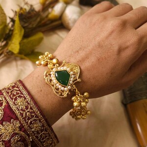 May include: Gold-toned bracelet featuring a central green gemstone, surrounded by small pearls. The ornate design includes delicate filigree and dangling gold beads. The bracelet is displayed on a wrist, with a maroon and gold embroidered sleeve visible.