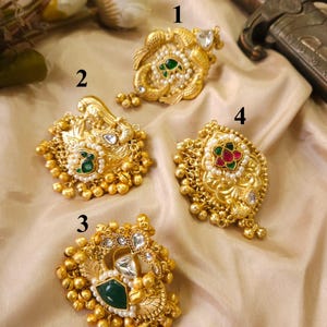 Puede incluir: Cuatro colgantes dorados ornamentados con diseños intrincados. Cada colgante presenta una piedra preciosa central en verde o rojo, rodeada de pequeñas perlas y cristales transparentes. Los colgantes se exhiben sobre una tela beige claro.