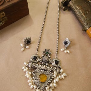 Puede incluir: Un conjunto de collar y pendientes plateados. El collar presenta un colgante triangular con una piedra amarilla, rodeado de perlas blancas y detalles cuadrados negros. Los pendientes tienen un dise&ntilde;o similar. Una caja de madera decorativa y una espada est&aacute;n en el fondo.