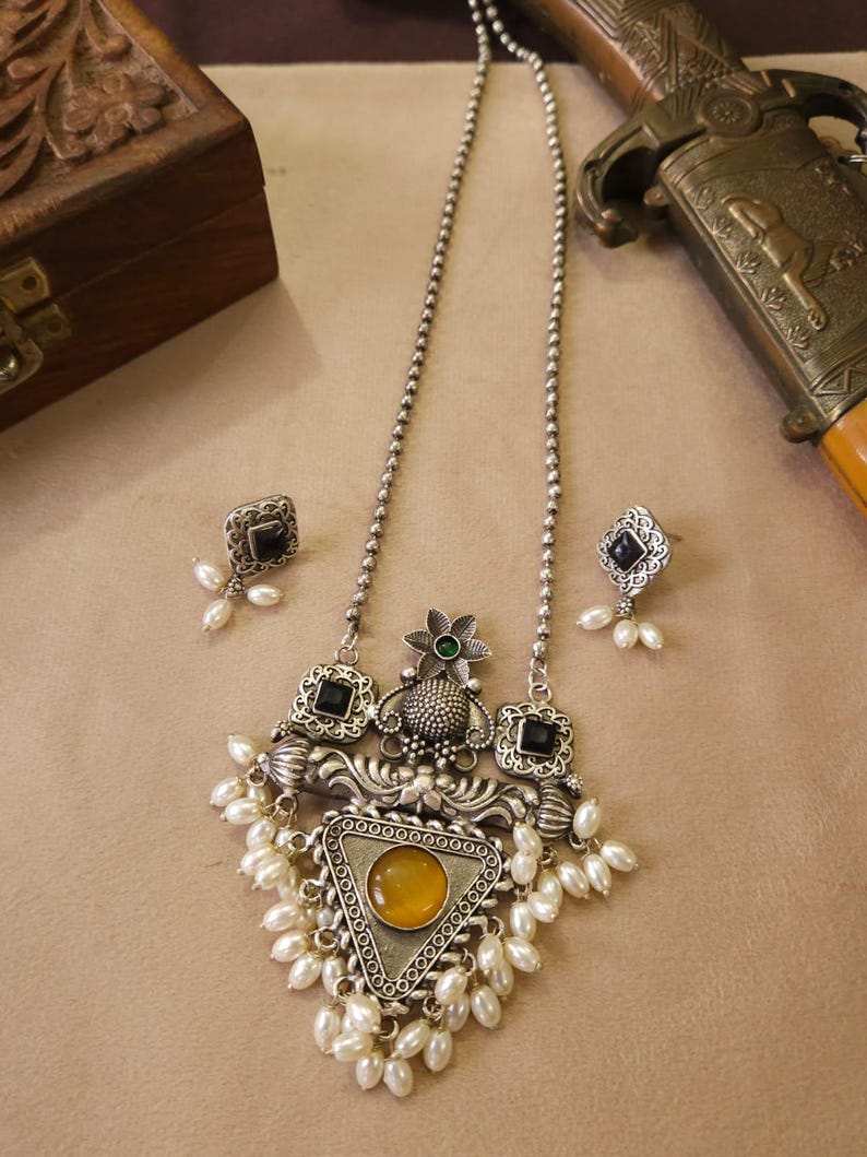 Puede incluir: Un conjunto de collar y pendientes plateados. El collar presenta un colgante triangular con una piedra amarilla, rodeado de perlas blancas y detalles cuadrados negros. Los pendientes tienen un dise&ntilde;o similar. Una caja de madera decorativa y una espada est&aacute;n en el fondo.