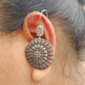 Puede incluir: Pendiente plateado "PIN BUGADI". El pendiente presenta un diseño grande, redondo y detallado con un centro floral y patrones intrincados. El pendiente se sujeta a la oreja con un gancho y un alfiler. El texto "Silver Replica" y "2056-A" son visibles.