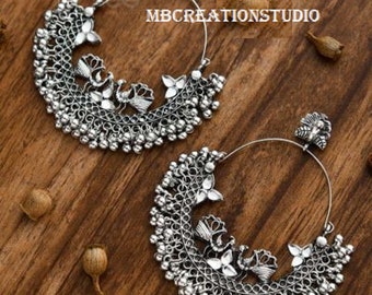 May include: A pair of silver hoop earrings with a peacock design and a beaded fringe. The earrings are on a wooden surface.