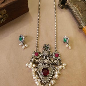 Puede incluir: Juego de collar y pendientes plateados. El collar presenta un colgante con una piedra roja triangular, rodeada de perlas blancas. Los pendientes tienen piedras cuadradas verdes y peque&ntilde;as perlas blancas. El conjunto tiene un dise&ntilde;o antiguo y ornamentado.
