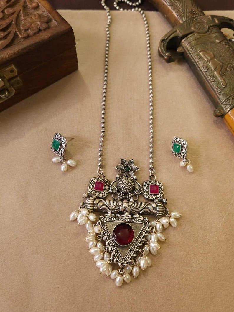 Puede incluir: Juego de collar y pendientes plateados. El collar presenta un colgante con una piedra roja triangular, rodeada de perlas blancas. Los pendientes tienen piedras cuadradas verdes y peque&ntilde;as perlas blancas. El conjunto tiene un dise&ntilde;o antiguo y ornamentado.