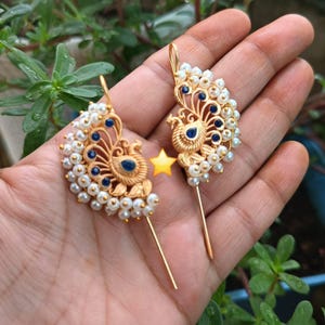 Op de afbeelding: Een paar goudkleurige pauwoorbellen. Elke oorbel heeft een pauwontwerp met blauwe accenten en kleine witte parels. De oorbellen hebben een lange, dunne pin voor inbrengen in het oor.