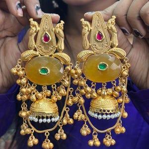 May include: Gold-toned earrings with intricate detailing. Each earring features a red and green gemstone, a large yellow stone, and dangling gold beads. The design includes bird motifs and bell-shaped elements, creating a luxurious look.