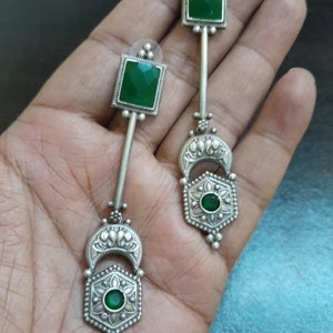 Op de afbeelding: Een paar zilveren oorhangers met groene edelsteen accenten. De oorhangers hebben een zeshoekig ontwerp met een kleinere groene edelsteen in het midden en een halve maanvorm erboven.