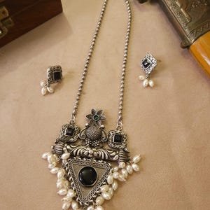 Puede incluir: Un conjunto de collar y pendientes plateados. El collar presenta un colgante triangular con una piedra negra y perlas blancas. Los pendientes tienen un dise&ntilde;o cuadrado con piedras negras y peque&ntilde;as perlas. Una caja decorativa y una espada est&aacute;n en el fondo.