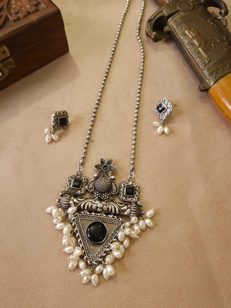 Puede incluir: Un conjunto de collar y pendientes plateados. El collar presenta un colgante triangular con una piedra negra y perlas blancas. Los pendientes tienen un dise&ntilde;o cuadrado con piedras negras y peque&ntilde;as perlas. Una caja decorativa y una espada est&aacute;n en el fondo.