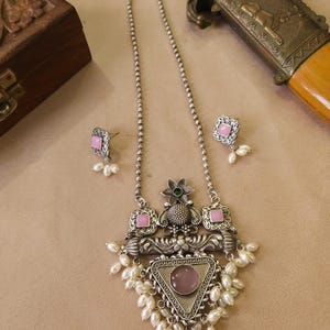 Puede incluir: Un conjunto de collar y pendientes plateados. El collar presenta un colgante con una piedra rosa, enmarcado por un tri&aacute;ngulo y adornado con peque&ntilde;as perlas blancas. Los pendientes tienen un dise&ntilde;o similar. El conjunto se exhibe sobre una superficie neutra.