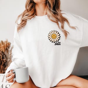 Könnte beinhalten: Weißes Sweatshirt mit einer Gänseblümchen-Grafik mit einem lächelnden Gesicht und dem Text "Sat Nam" in Schwarz. Das Gänseblümchen hat eine gelbe Mitte und einen schwarzen Umriss. Das Sweatshirt ist ein lässiger, bequemer Stil.