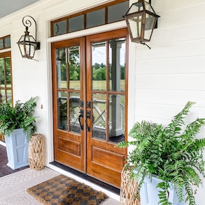 May include: Exterior view of a home's entrance featuring a pair of wooden doors with glass panels. The doors are flanked by ferns in light blue planters and decorative lanterns. A patterned doormat sits on a patterned rug.