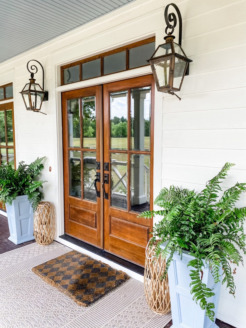 May include: Exterior view of a home's entrance featuring a pair of wooden doors with glass panels. The doors are flanked by ferns in light blue planters and decorative lanterns. A patterned doormat sits on a patterned rug.