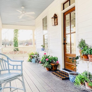 May include: A charming porch with a light blue ceiling and a white ceiling fan. The porch features a light blue wicker chair, potted plants, and a wooden door with glass panels. A welcome mat sits in front of the door.